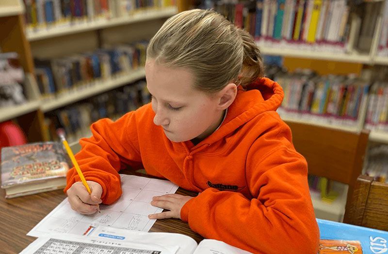 Girl in orange hoodie writing at a table in a library, holding a pencil, with books nearby. The Immanuel Difference | Immanuel Lutheran School | Belvidere, IL