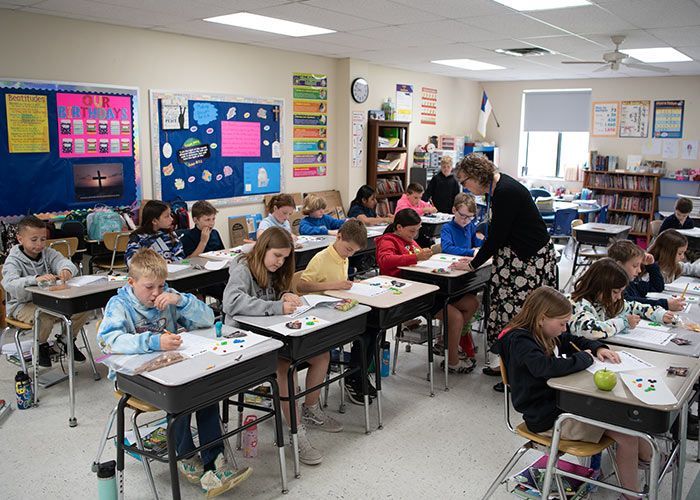 Students work at desks in a classroom; a teacher assists them.  Immanuel Lutheran School | Belvidere, IL