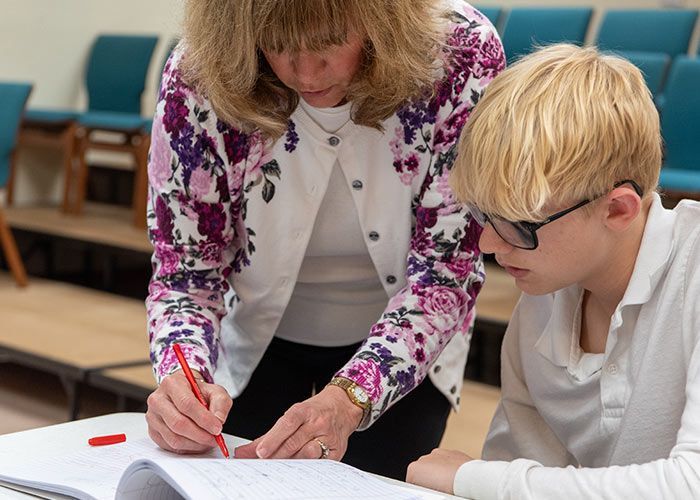 Woman points red pen at notebook while assisting a person wearing glasses. History | Immanuel Lutheran School | Belvidere, IL