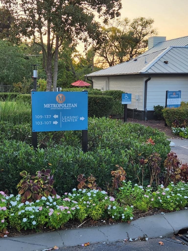 A blue sign is in the middle of a garden in front of a building.