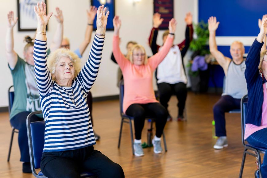 A group of seniors in chairs, raising arms during a seated exercise class in a bright room.