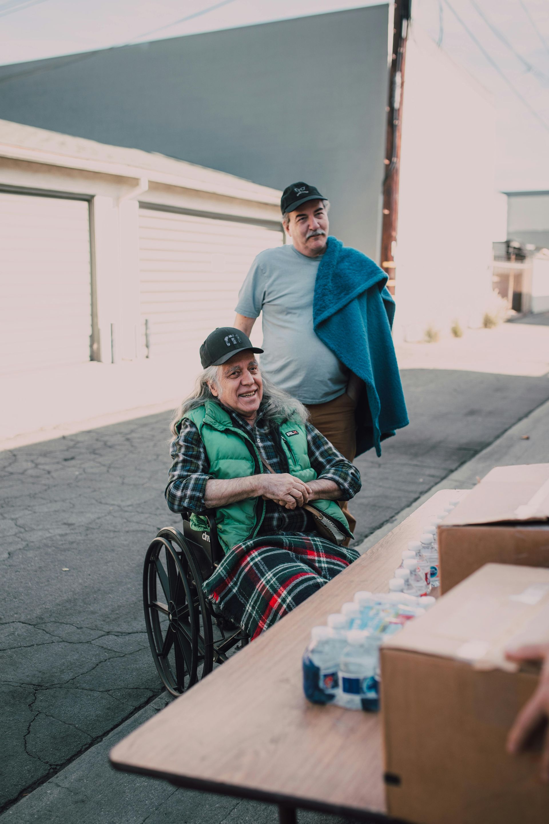 An elderly person in a wheelchair smiles, wearing a cap and plaid blanket. A man stands behind him, holding a blue blanket. They are outside at a table with boxes.