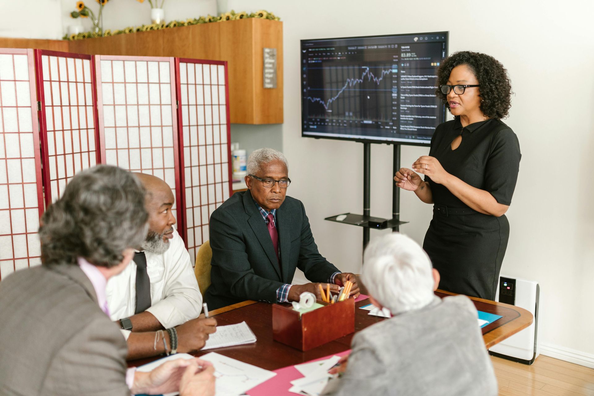 Woman in a black dress leading a business meeting, presenting data on a screen to four seated colleagues in a brightly lit room.