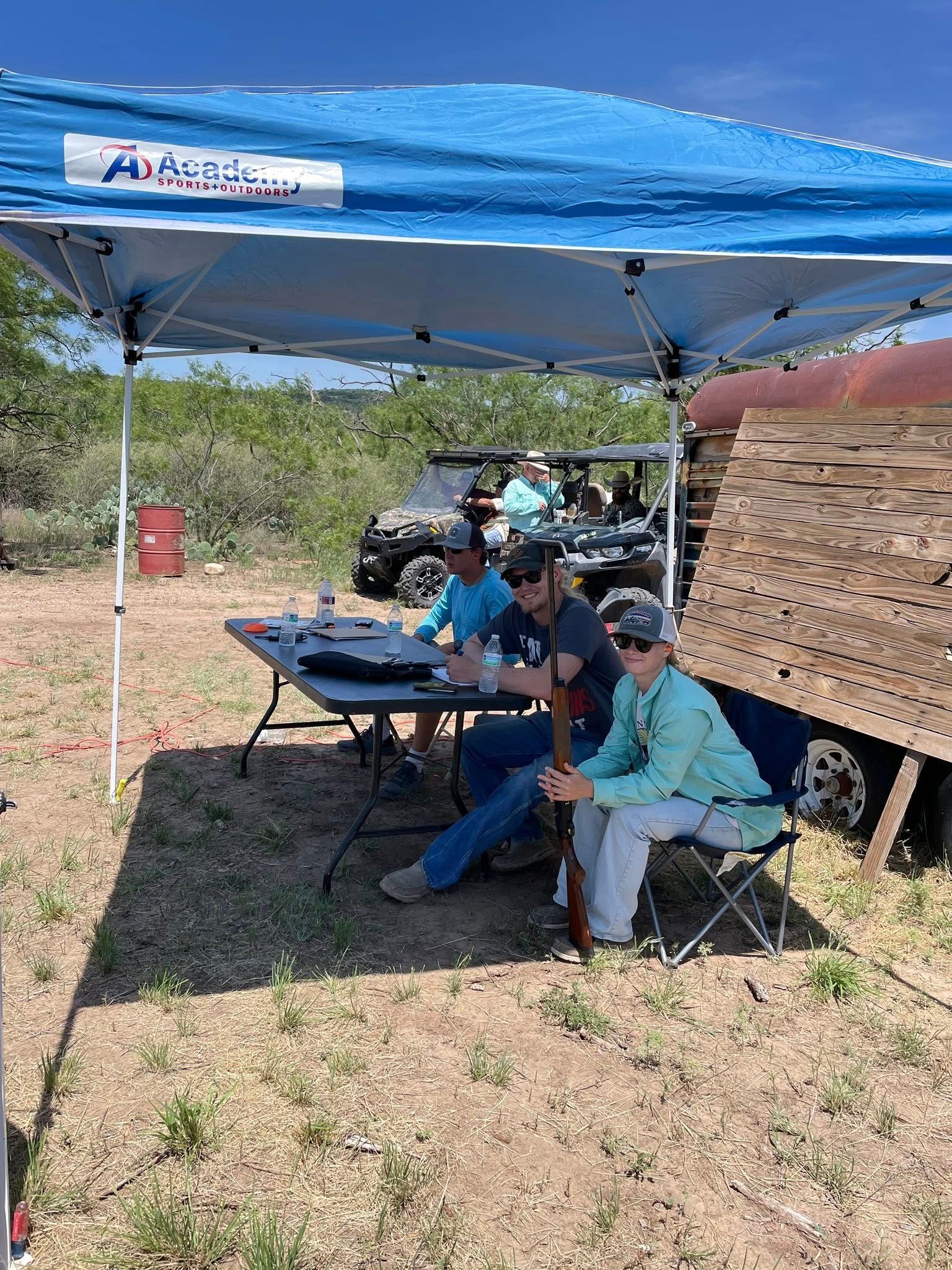 A group of people are sitting under a blue tent in a field.