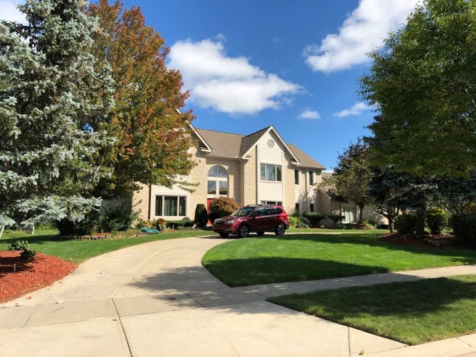 A two-story beige house with a red SUV parked in the driveway, under a partly cloudy blue sky.