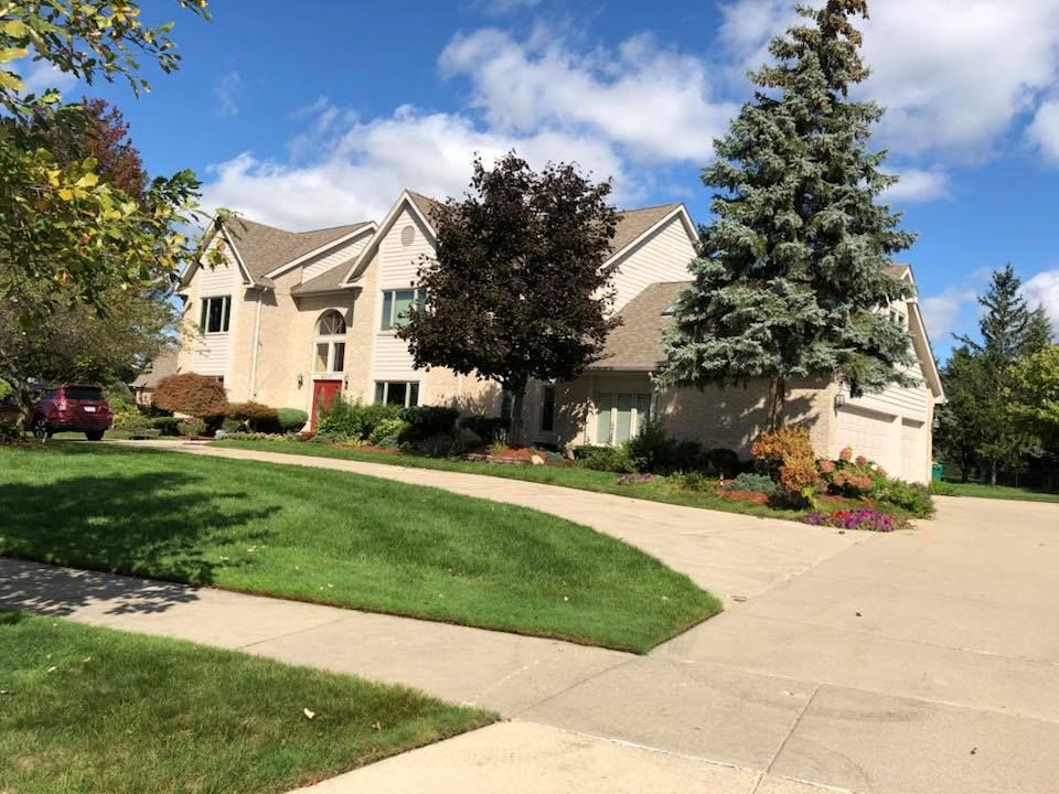 Large beige house with a circular driveway and green lawn under a blue sky with clouds.