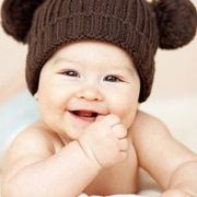 Smiling baby wearing a brown knit hat with two pom-poms, indoors.