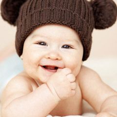 Smiling baby wearing a brown knit hat with two pom-poms, indoors.