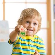 Young child with blonde hair smiles while holding a wooden block, wearing a striped shirt.