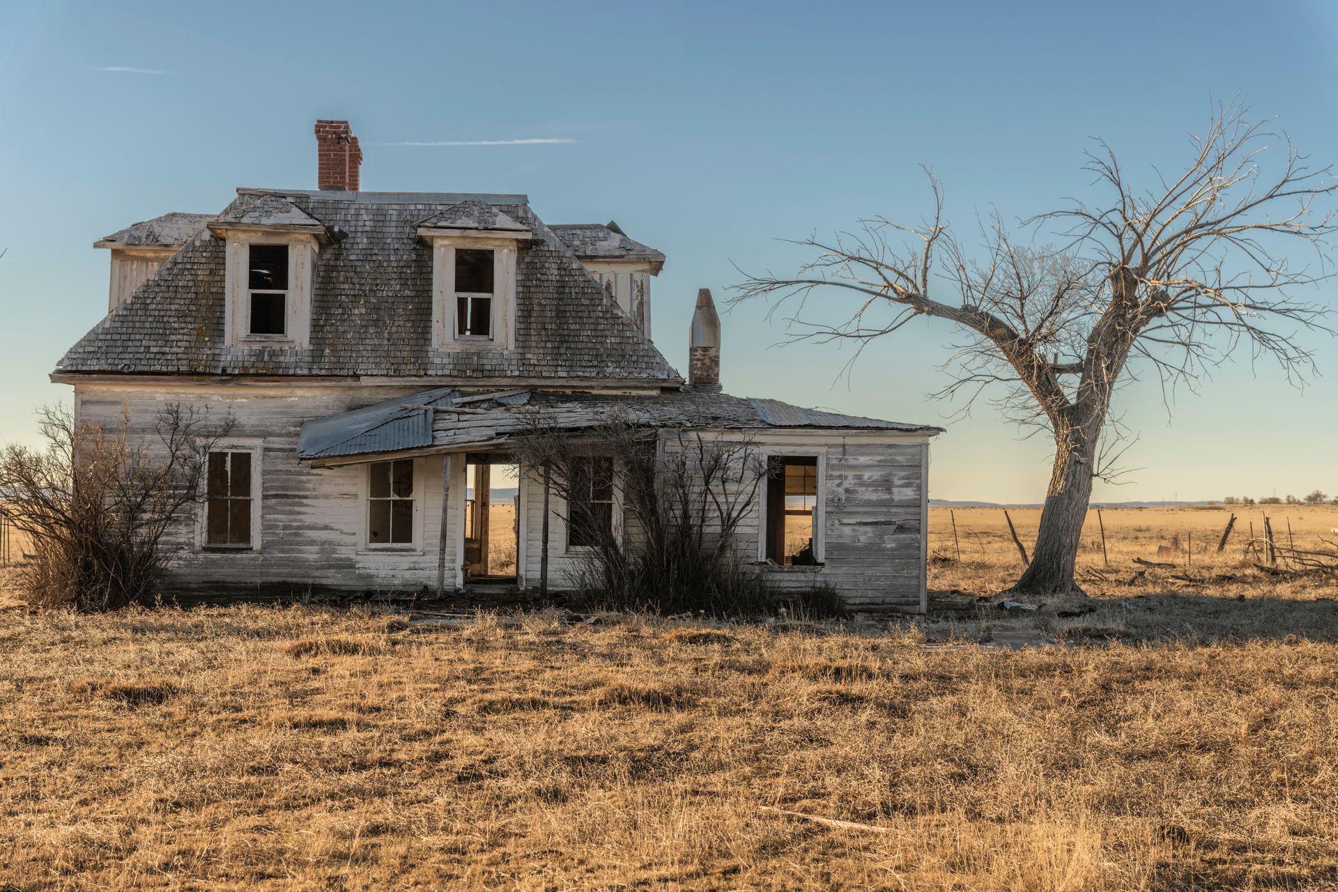 Dilapidated two-story farmhouse with peeling paint and bare tree in a dry, open field under a blue sky.