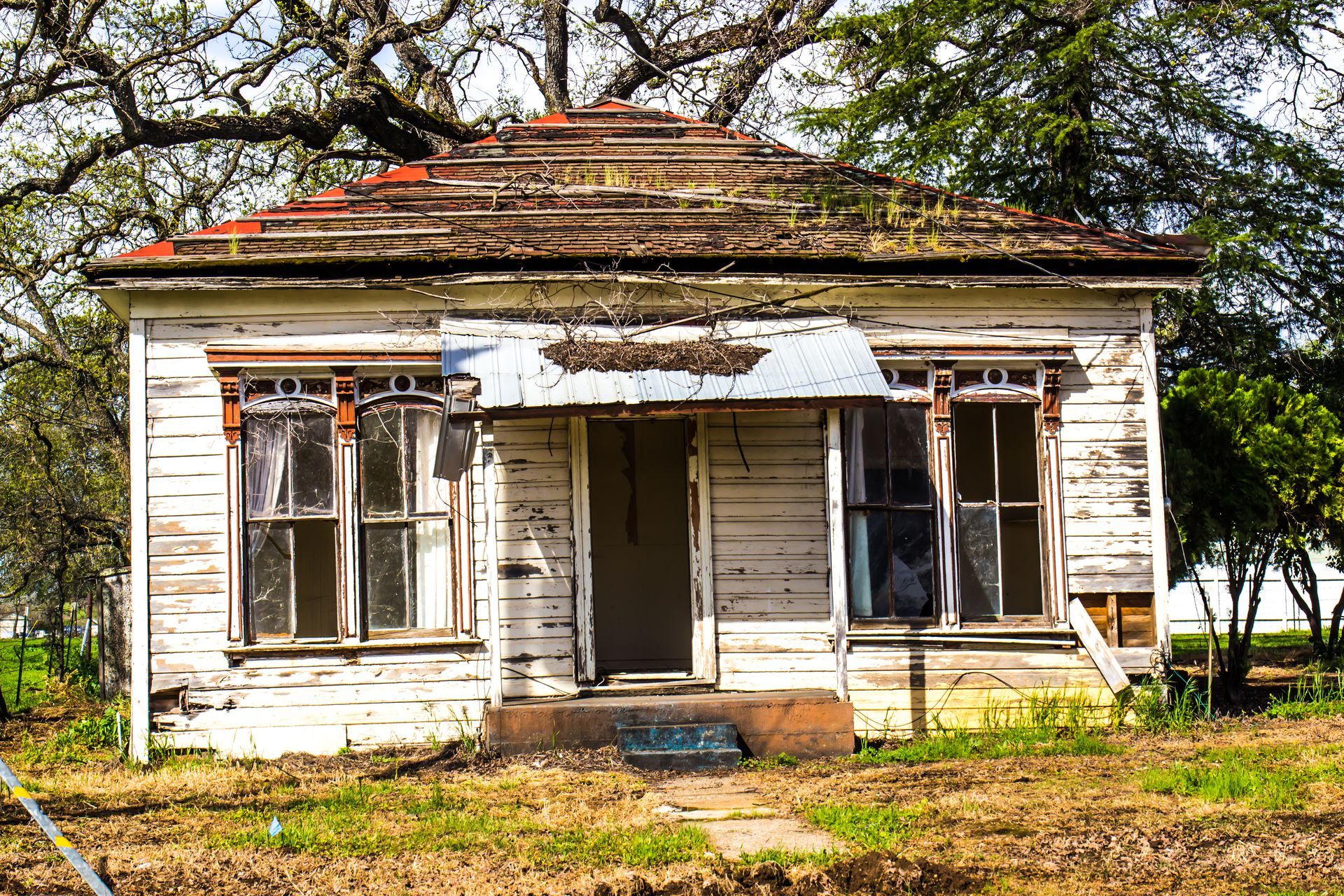 Dilapidated white wooden house with peeling paint, broken windows, and overgrown vegetation.