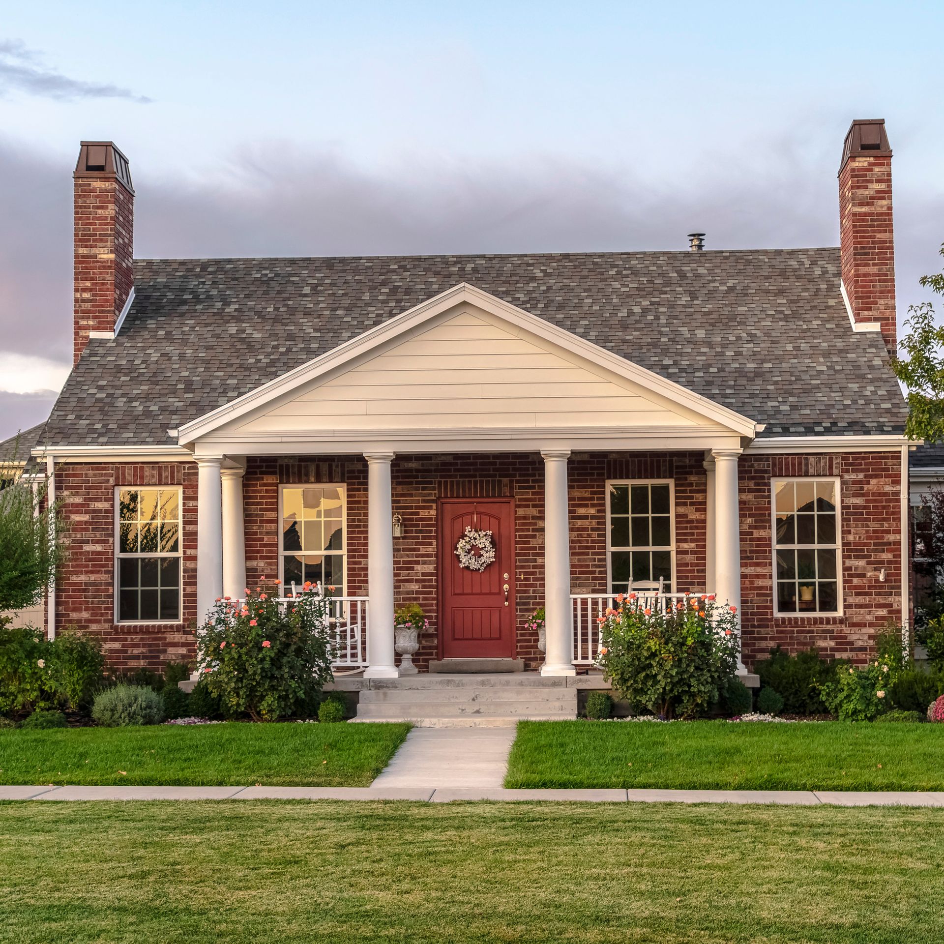 Brick house with a red door, white porch, and chimneys on a green lawn.