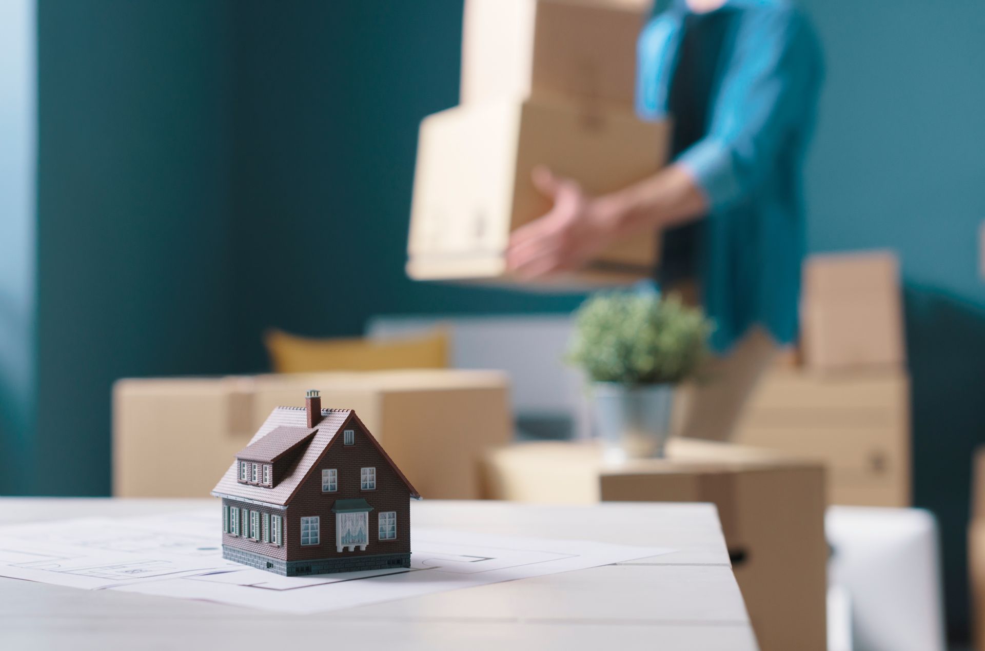 Model house on table; person carries moving boxes in new home.