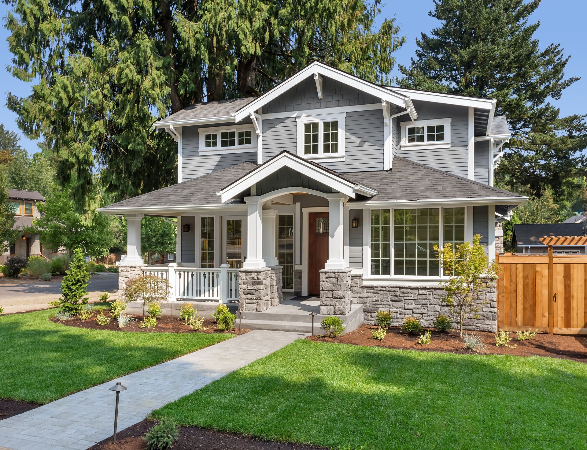 Gray two-story house with stone accents, a porch, and a walkway leading to the front door. Lush green lawn and trees.