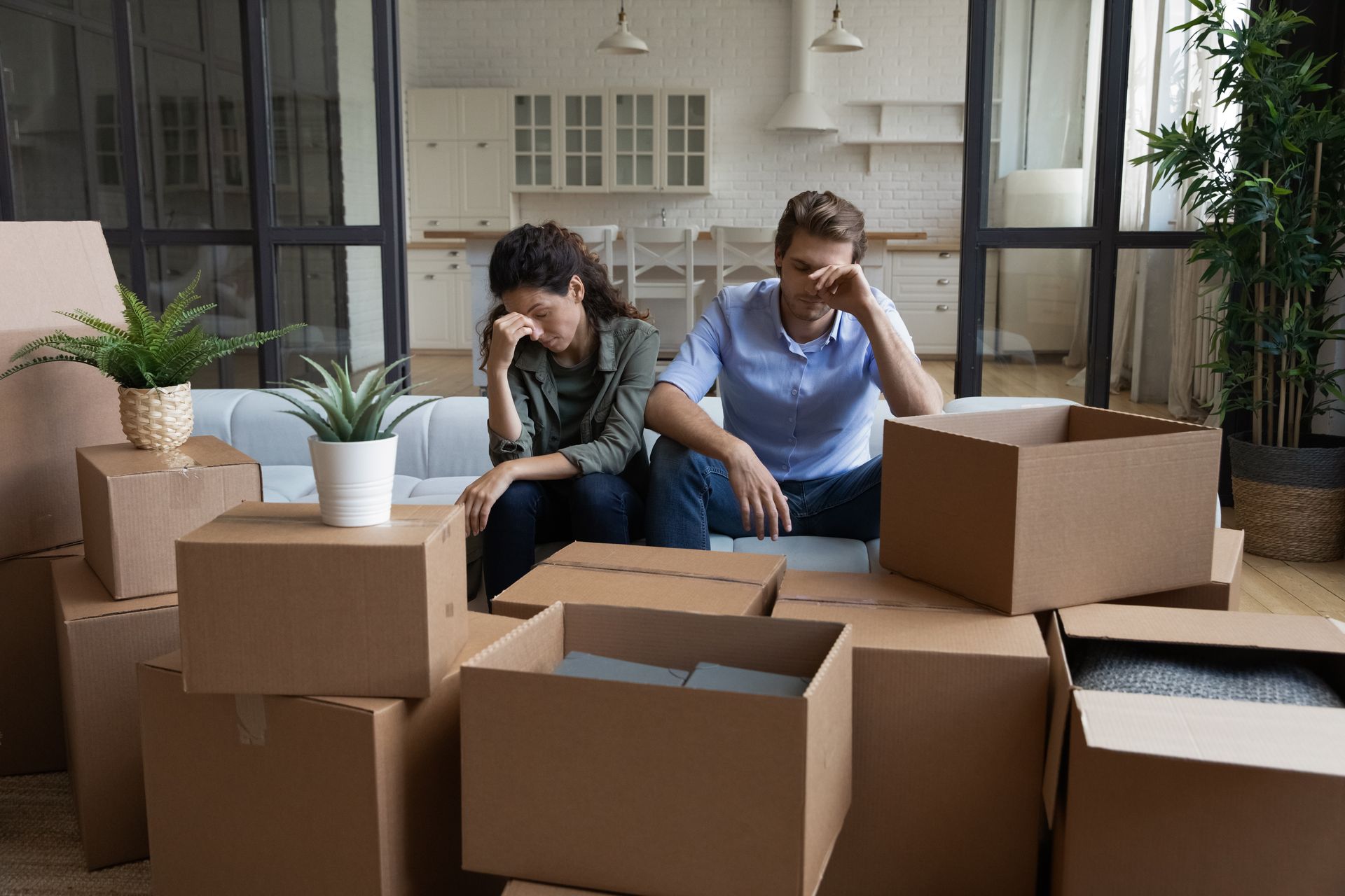 Couple sits amidst moving boxes, looking stressed; indoor setting.