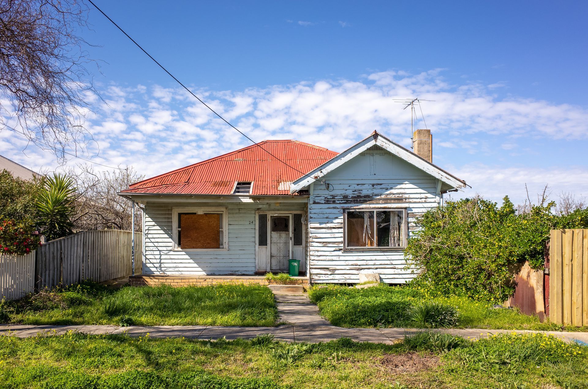 Dilapidated blue house with a red roof, surrounded by overgrown grass, on a sunny day.