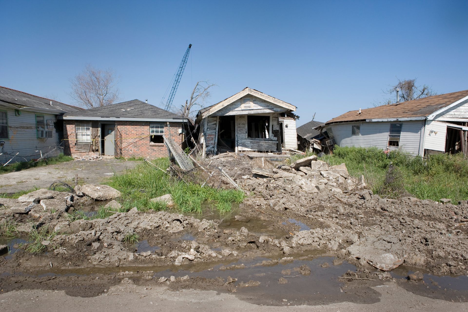 Homes damaged, debris-filled yards, and muddy street; potential aftermath of a natural disaster.