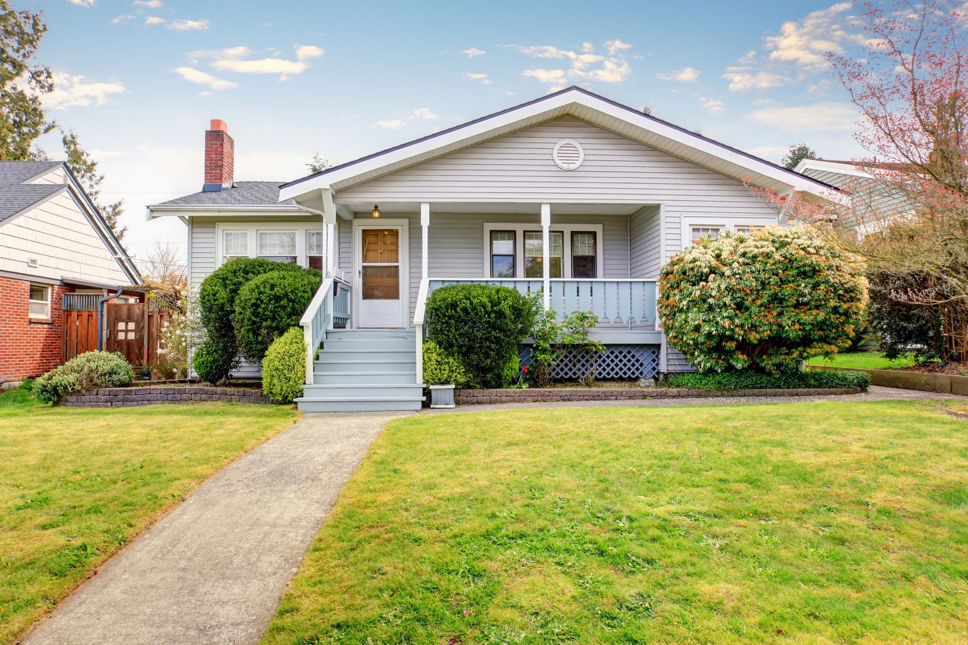 A light grey bungalow with a porch, chimney, and a green lawn with a sidewalk.