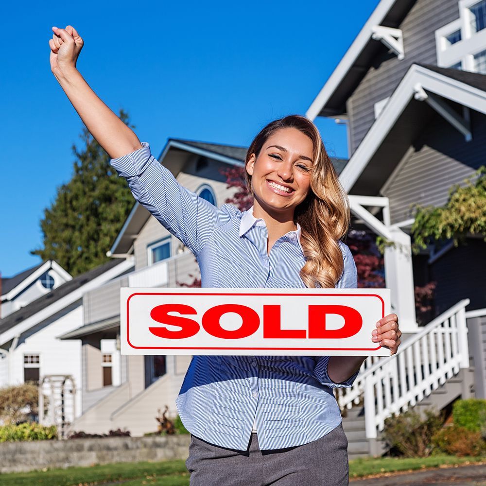 A smiling woman with a red SOLD sign, standing in front of a row of suburban houses with her right a