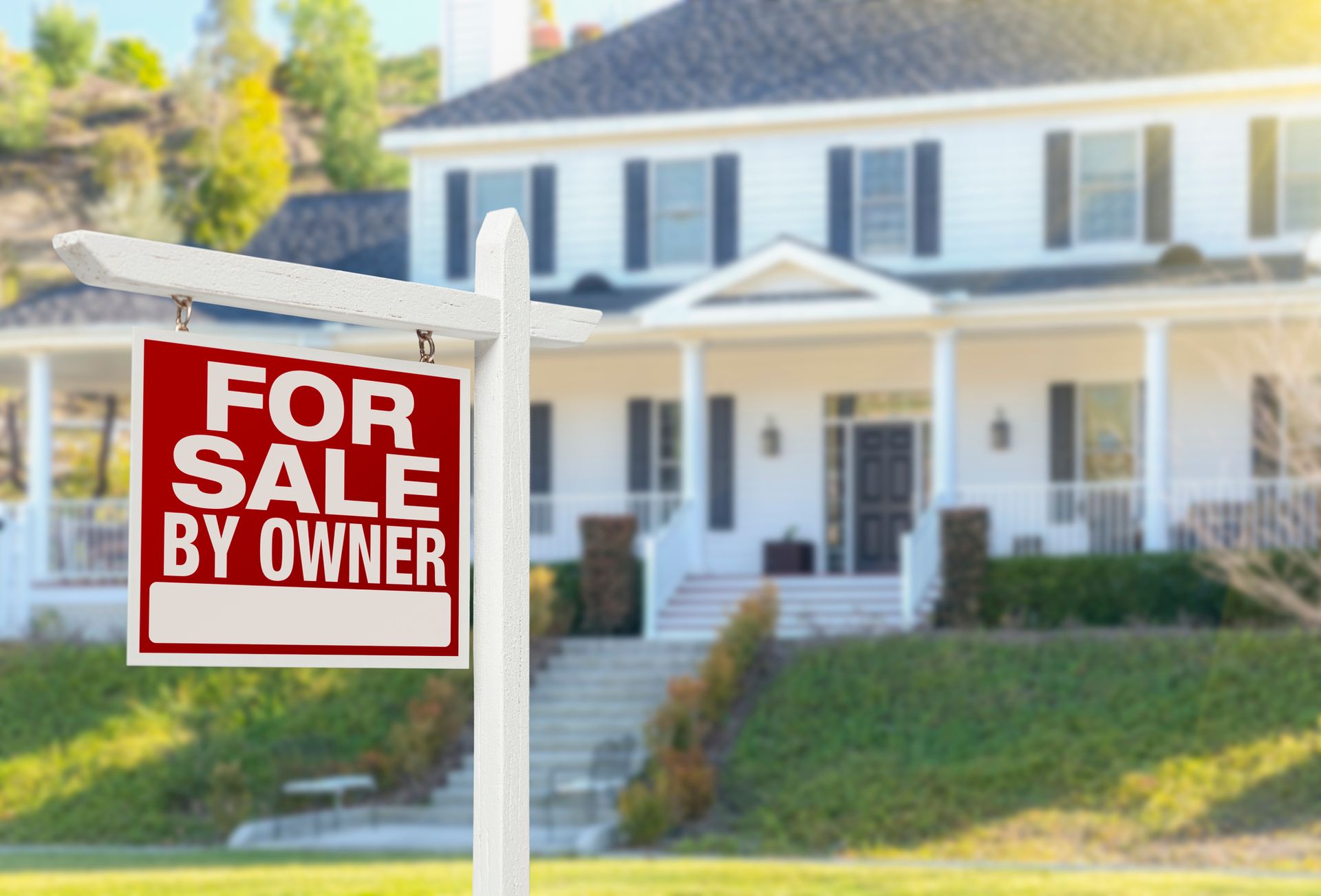 White house with porch and “For Sale by Owner” sign on lawn.