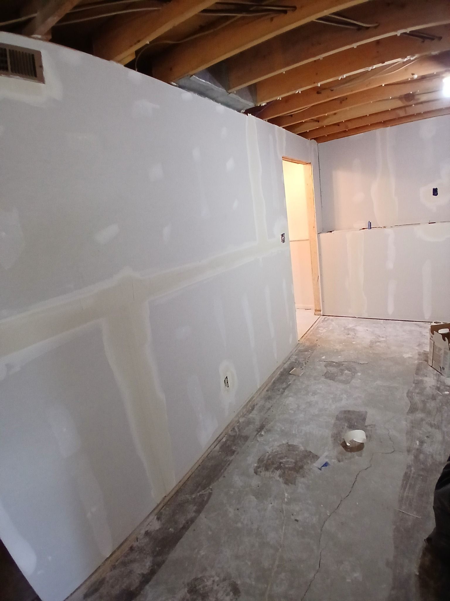 Interior view of a room under construction, drywall installation is in progress; bare concrete floor, wooden ceiling beams.