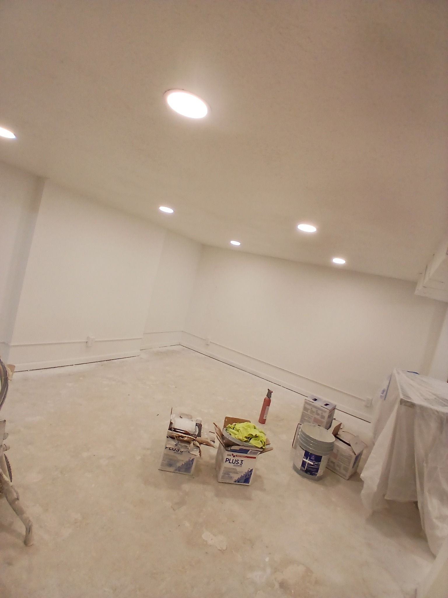 White-walled room under construction with recessed lighting and paint supplies on the floor.