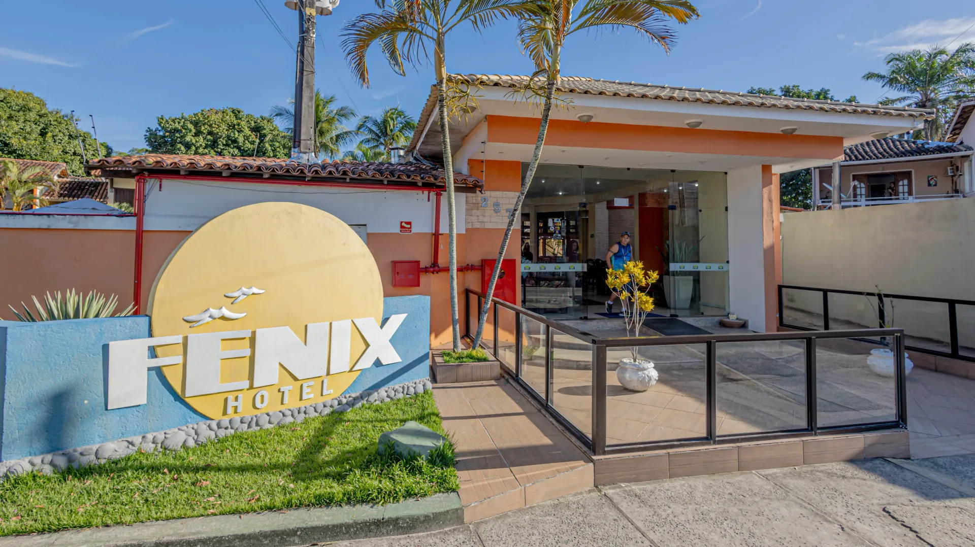 Placa e entrada do Hotel Fenix, com uma pequena passarela e portas de vidro. Dia ensolarado, cores azul e laranja.