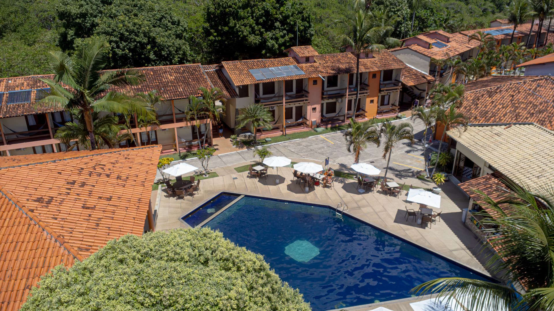 Vista aérea de um resort com edifícios, piscina e guarda-sóis. Árvores verdes rodeiam as estruturas. Hotel Fênix