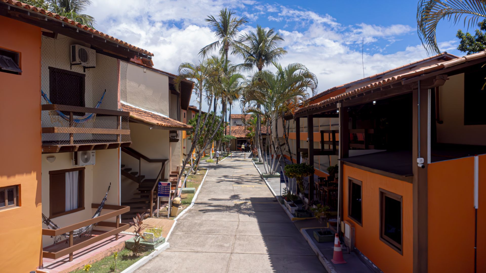 Um caminho pavimentado entre prédios de dois andares, palmeiras e um céu azul. Hotel Fênix