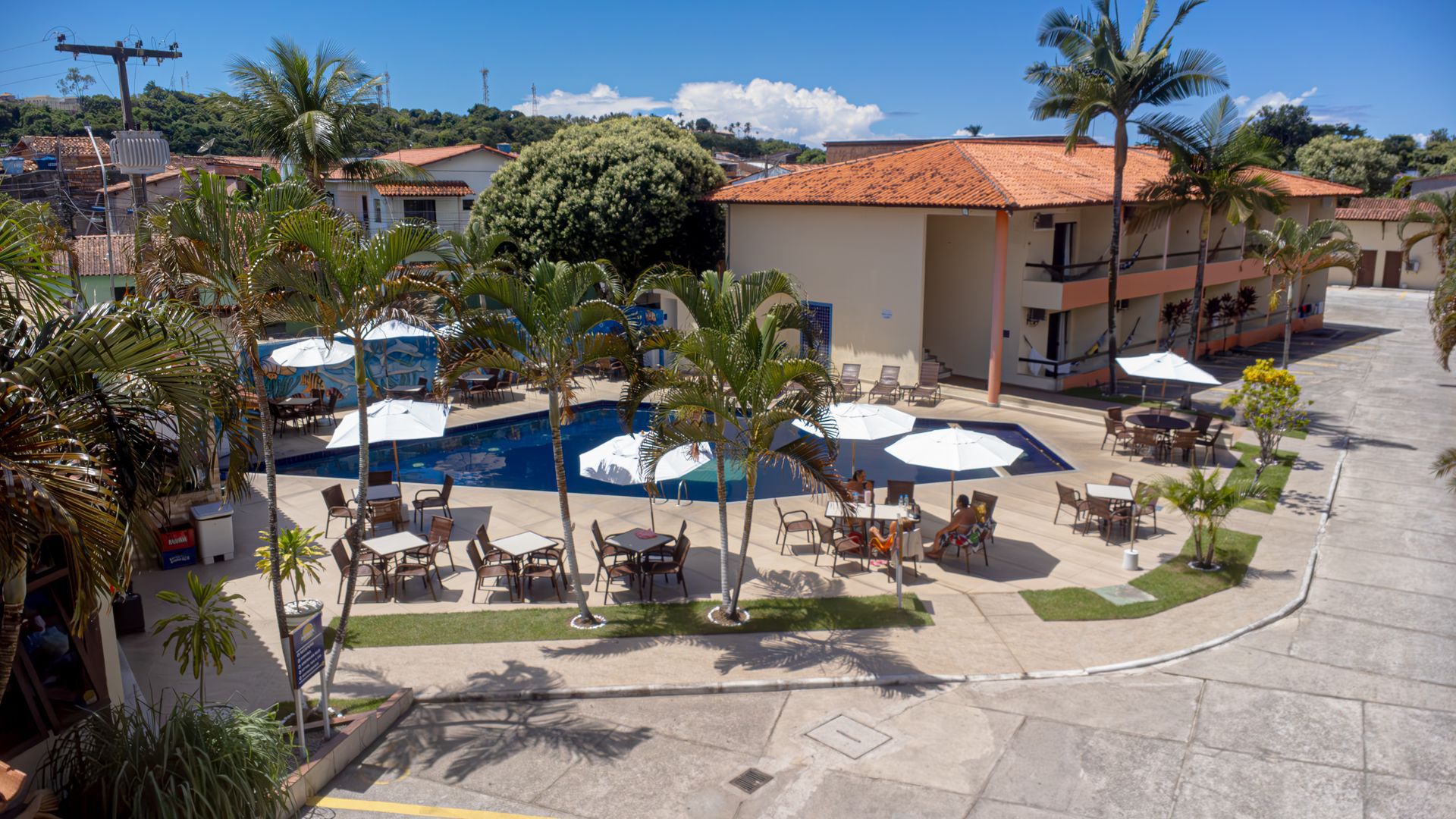 Exterior do hotel com piscina, mesas, guarda-sóis e palmeiras sob um céu azul. Hotel Fênix