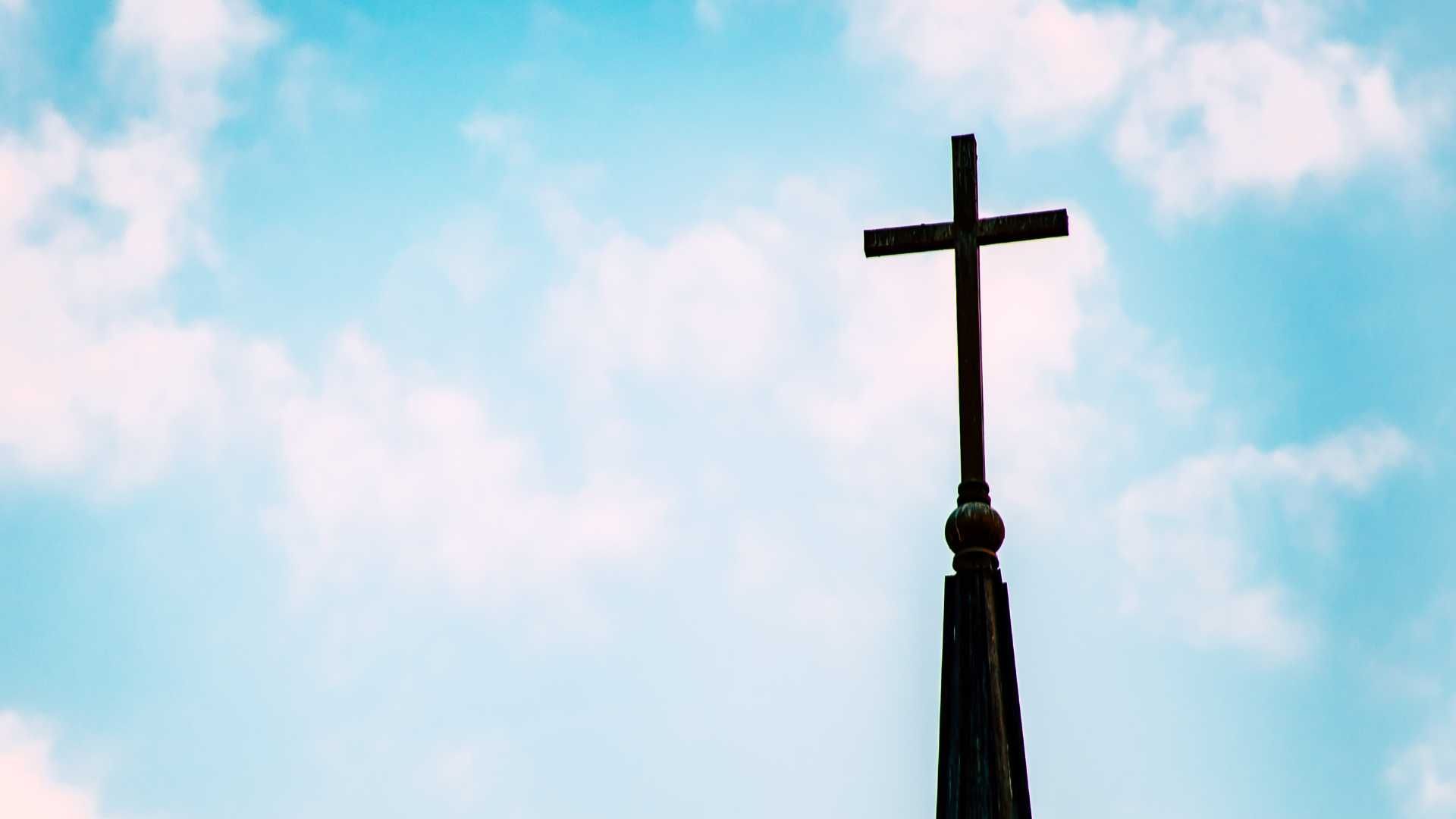 A church steeple with a cross on top of it against a cloudy blue sky.
