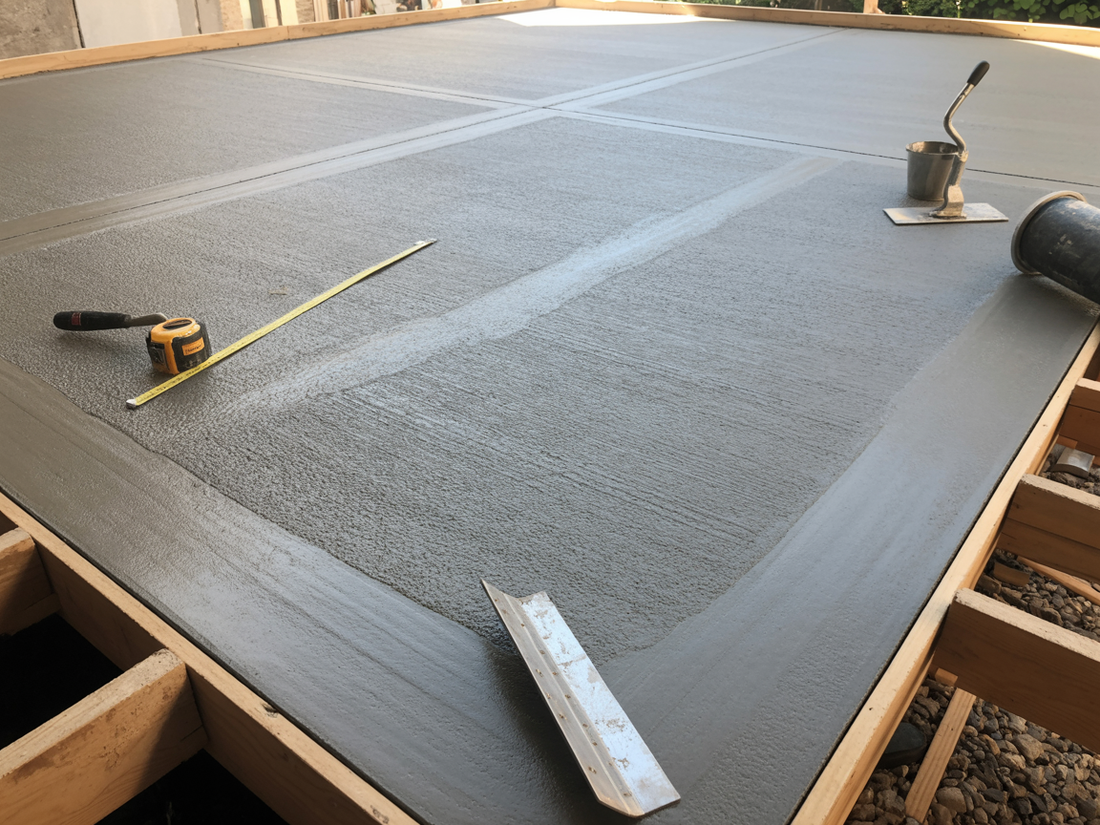Gray epoxy floor being applied in a large room, with tools and a trowel on the surface.