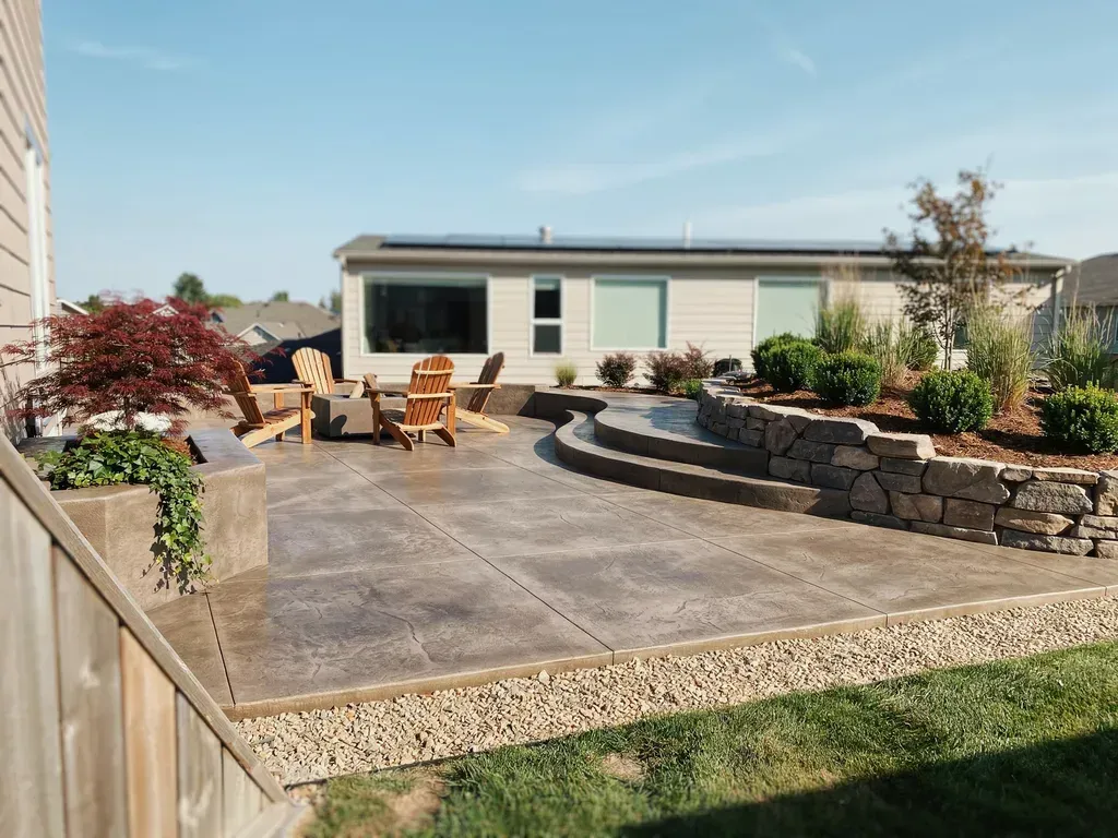 Backyard patio with concrete seating area, low stone wall, and modern house under a clear sky