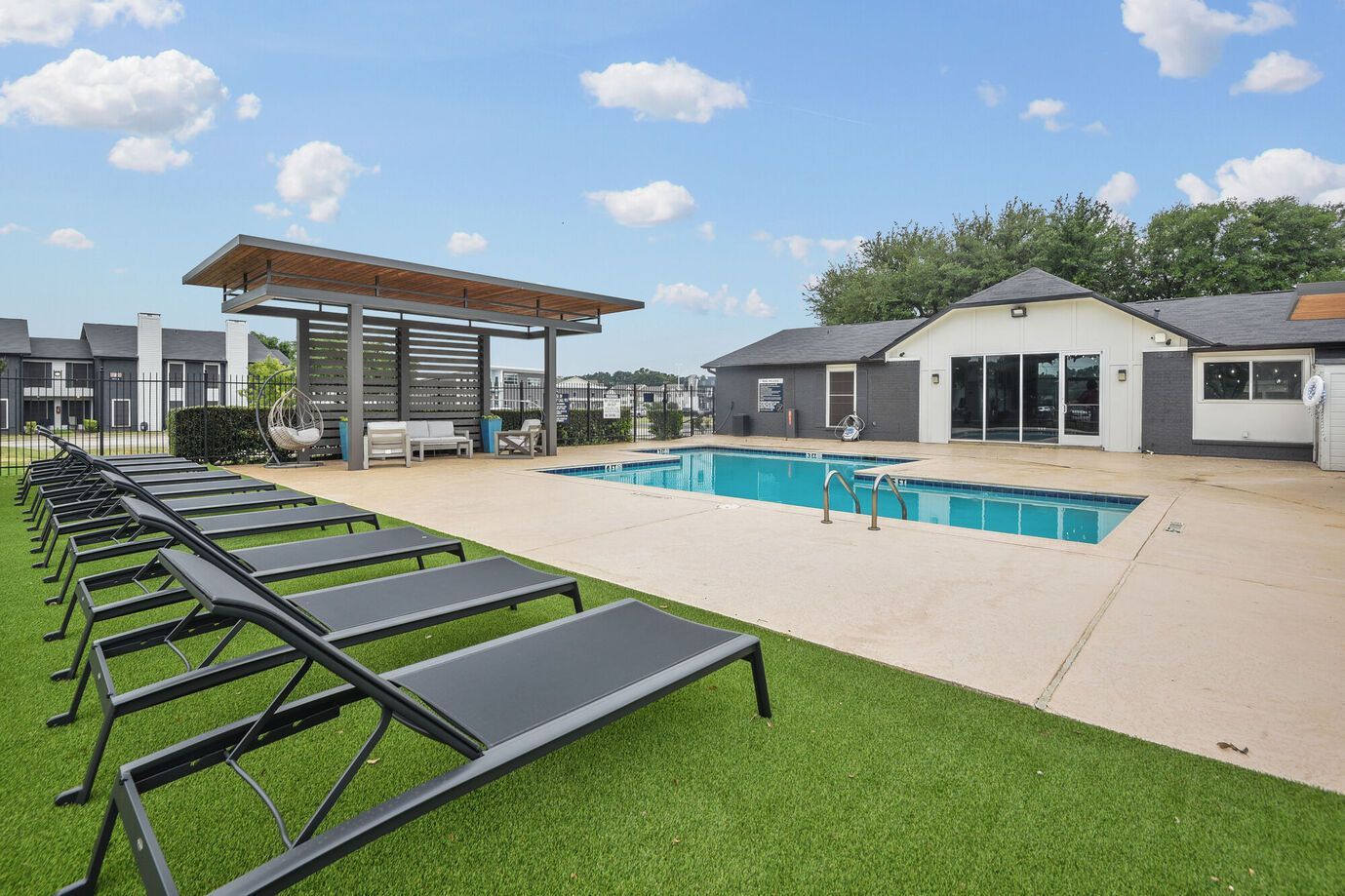 Pool area with lounge chairs on artificial turf, a pool, and a covered seating area under a blue sky.