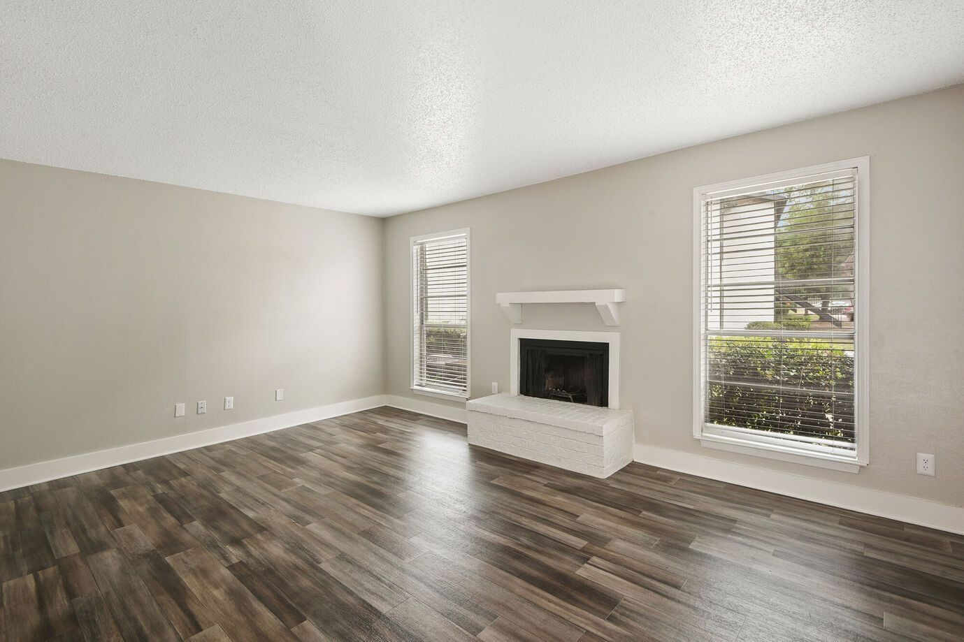 Empty living room with fireplace, two windows, and wood flooring. Walls are beige.