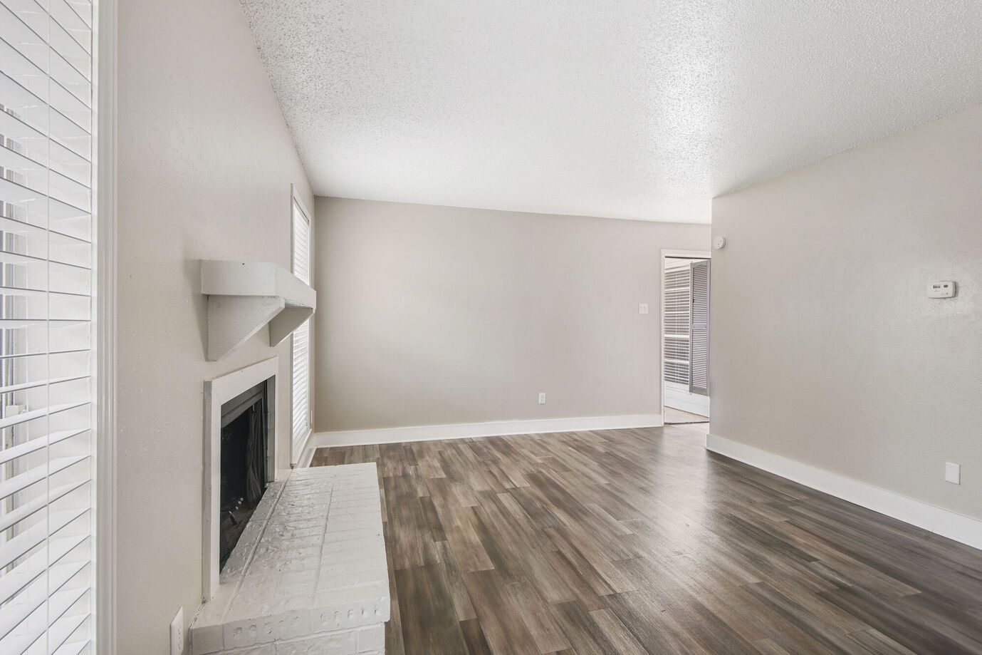 Empty living room with gray walls, wood flooring, fireplace, and white blinds.