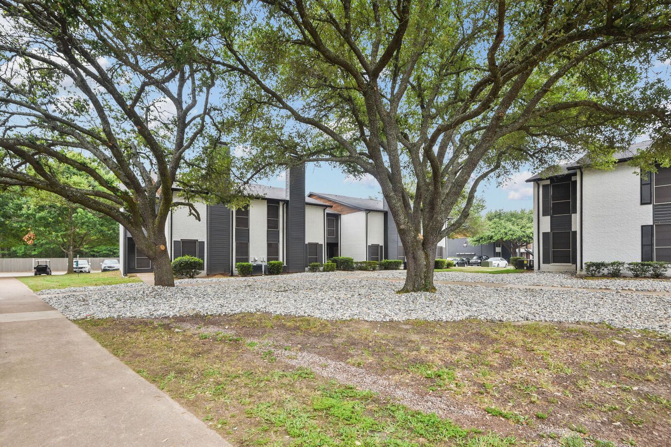 Apartment buildings with white exteriors and black accents, set among large trees and a rock garden.