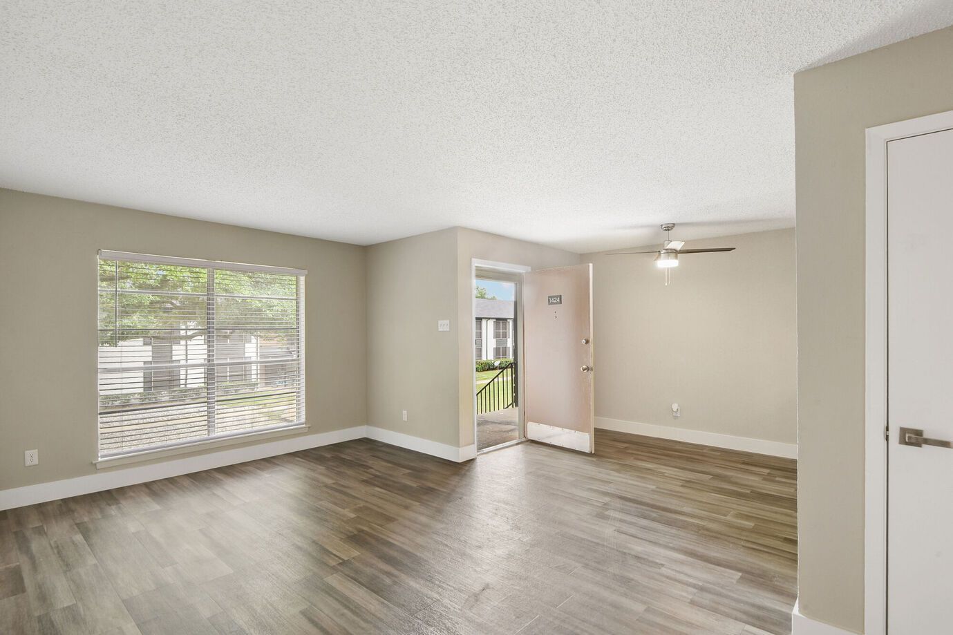 Empty living room with wood-look floors, light-colored walls, and a window overlooking greenery.