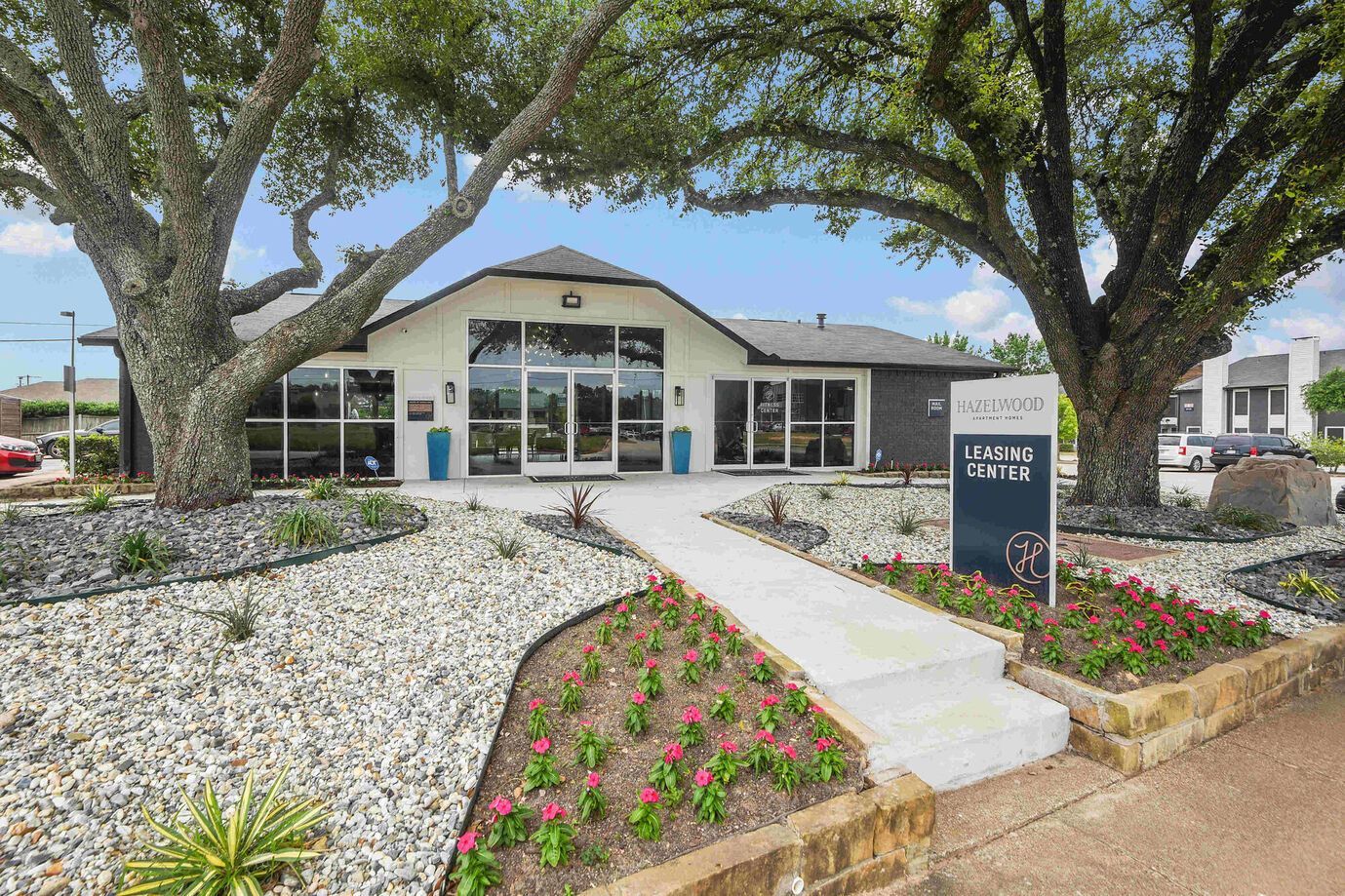 White building with glass doors under large trees, pathway lined with rocks and flowers.