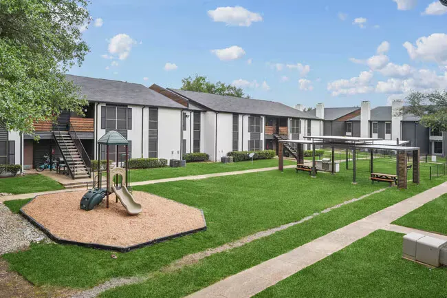 Apartment complex with a playground and picnic area on a green lawn, under a partly cloudy sky.