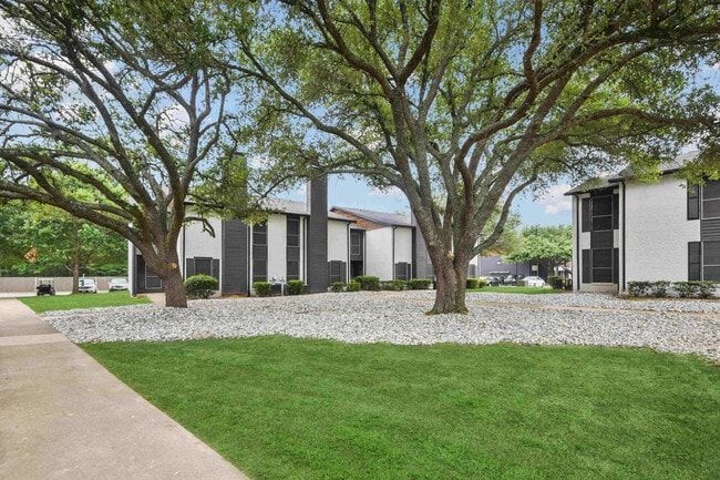 Apartment buildings with trees and gravel beds, sidewalk, green lawn, overcast sky.