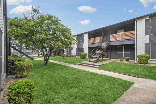Apartment complex exterior with grass, walkway, and tree under a blue sky.