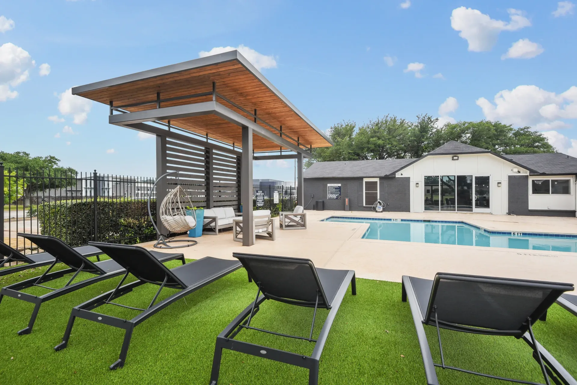 Poolside lounge area with sunbeds, canopy, pool, and building on a sunny day.