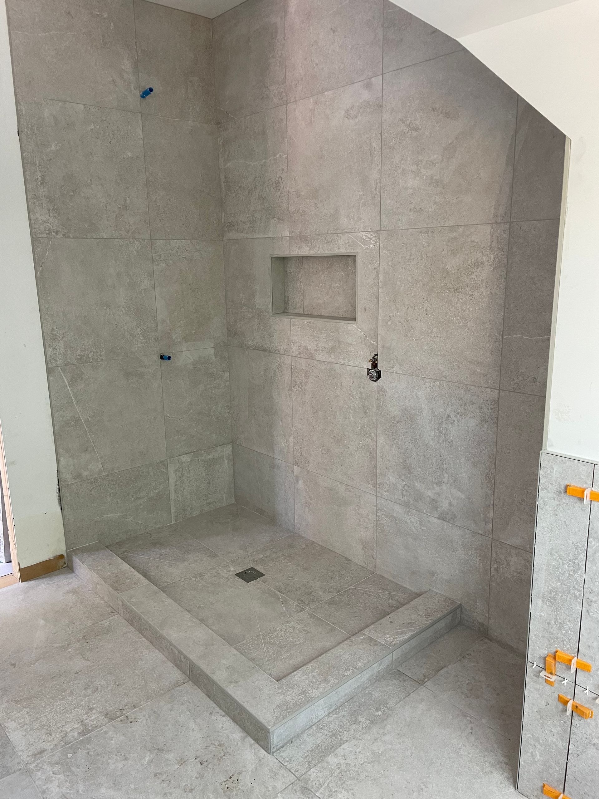 Cabine de douche en construction, avec murs carrelés gris, étagère encastrée et rebord.