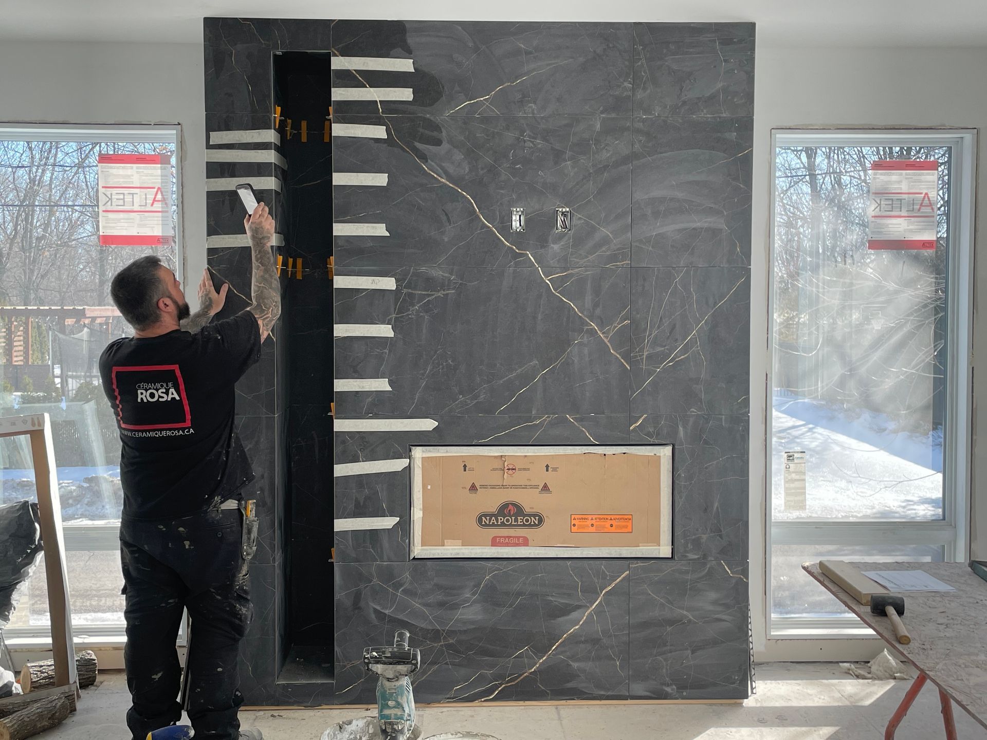 Un homme pose des carreaux de marbre foncé sur une cheminée dans une pièce avec fenêtres ; des travaux électriques sont visibles.