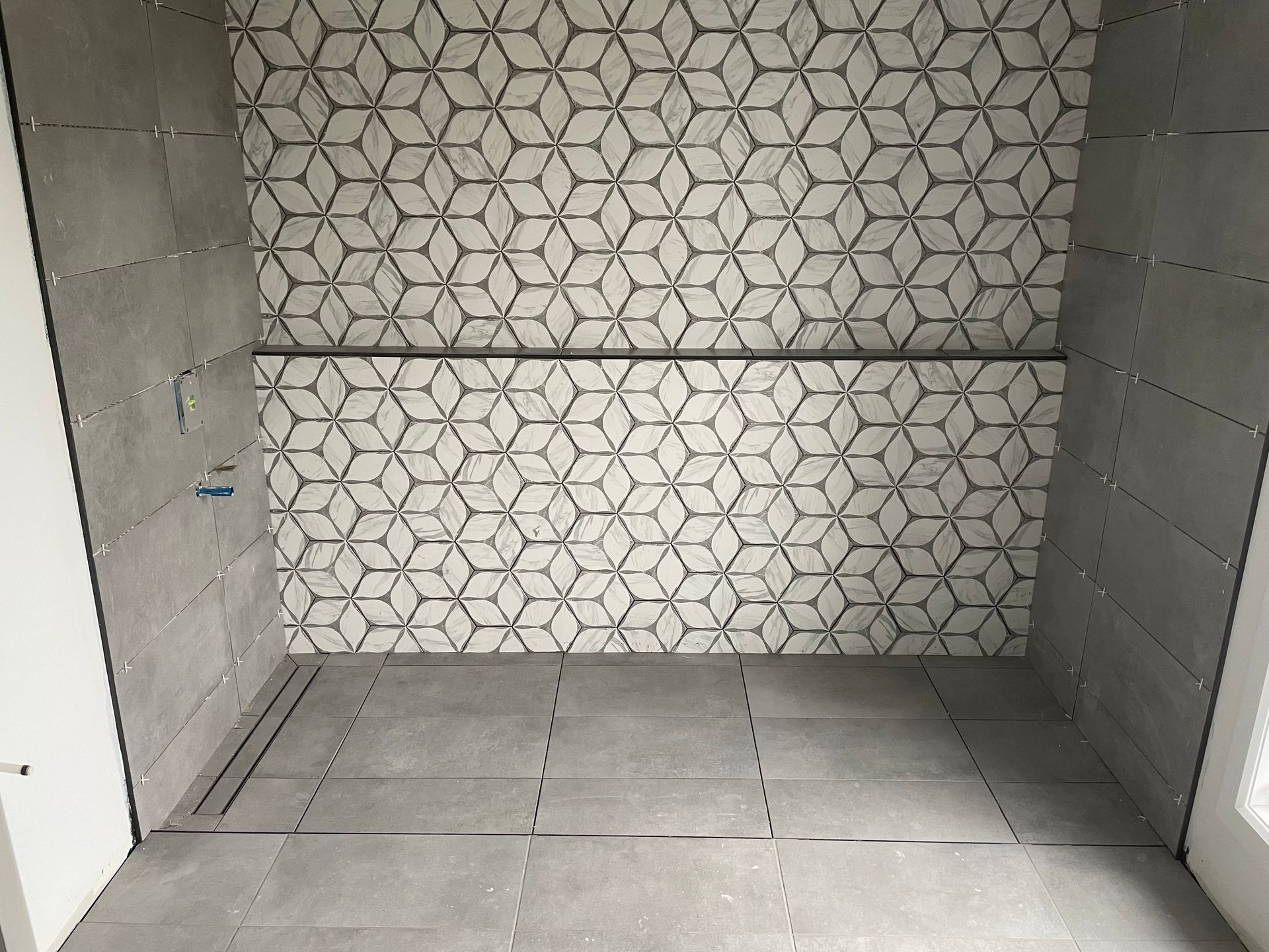 Salle de bains en construction avec carrelage gris au sol et aux murs, et carreaux décoratifs à motifs sur le mur.