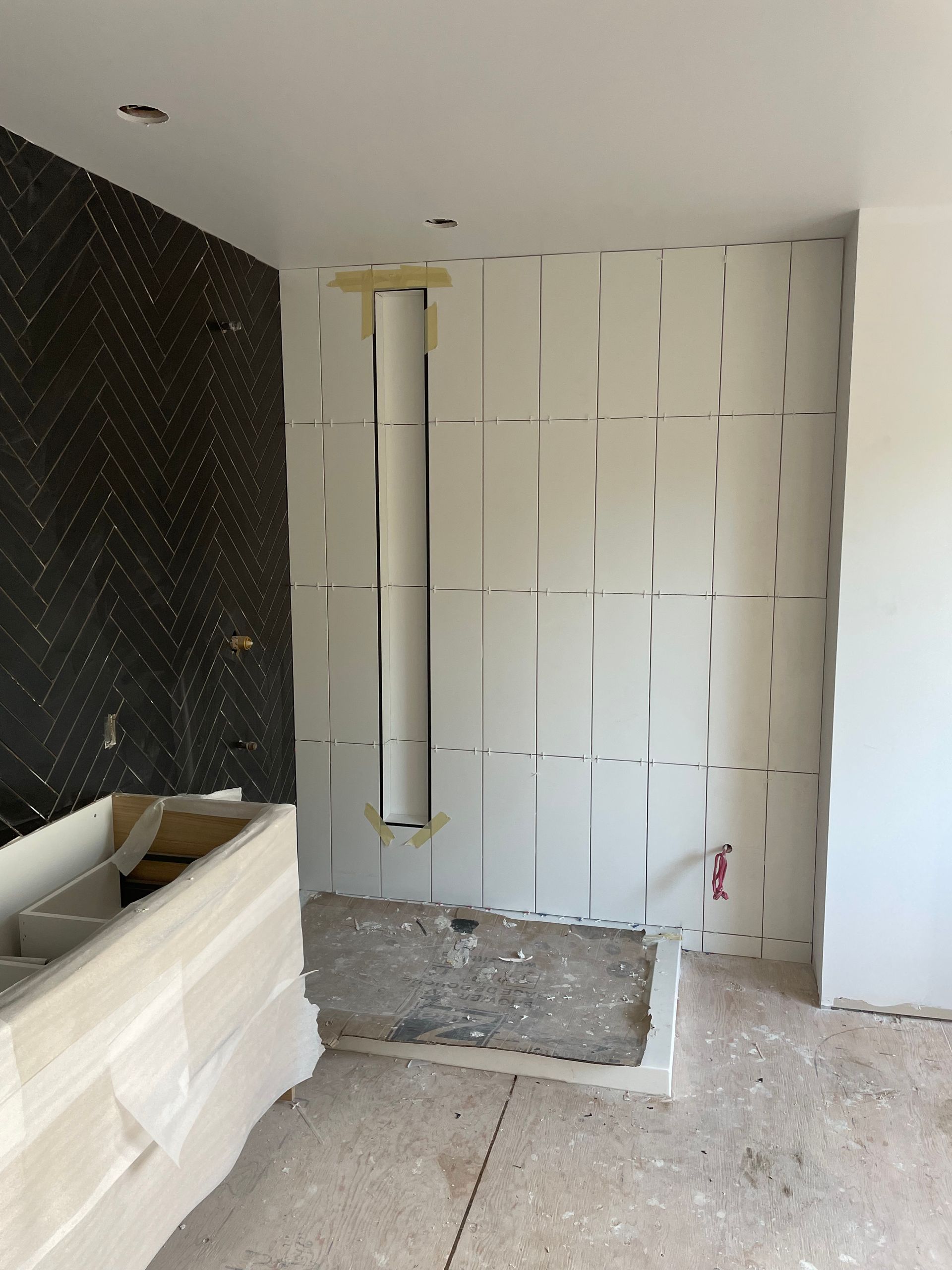 Chantier : mur carrelé de carreaux blancs texturés, mur d’accent en chevrons noirs, sol en béton brut et étagère intégrée.