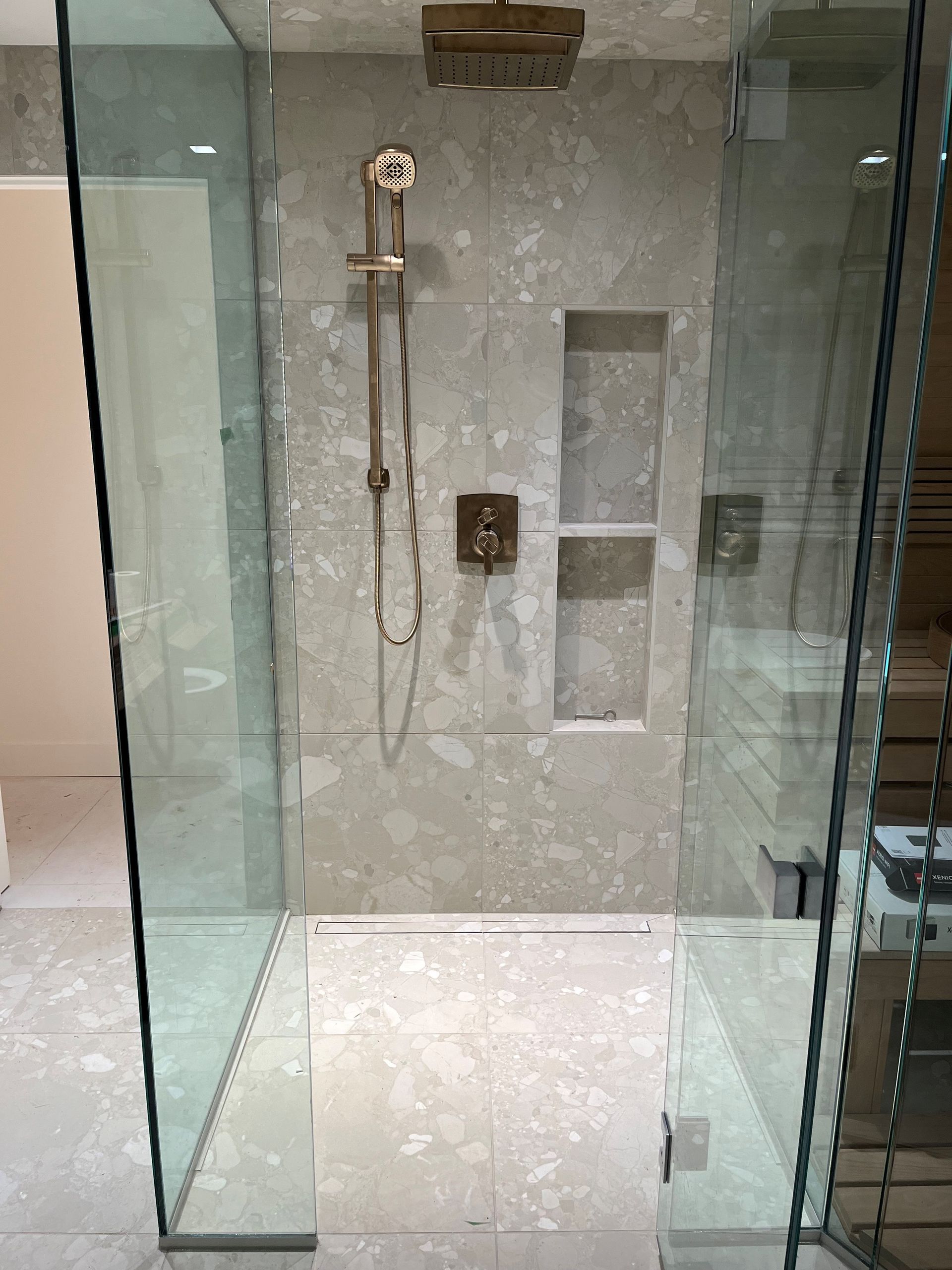 Douche vitrée avec robinetterie dorée et murs carrelés. Étagère encastrée. Sol carrelé gris.