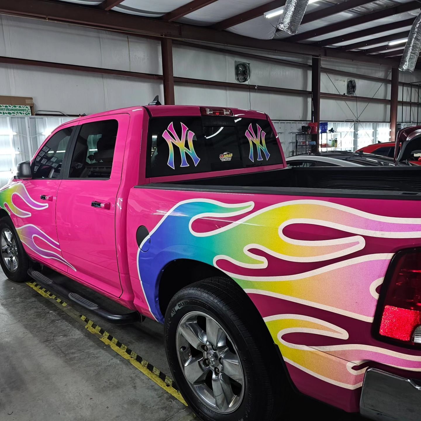 A pink truck with rainbow flames painted on it is parked in a garage.