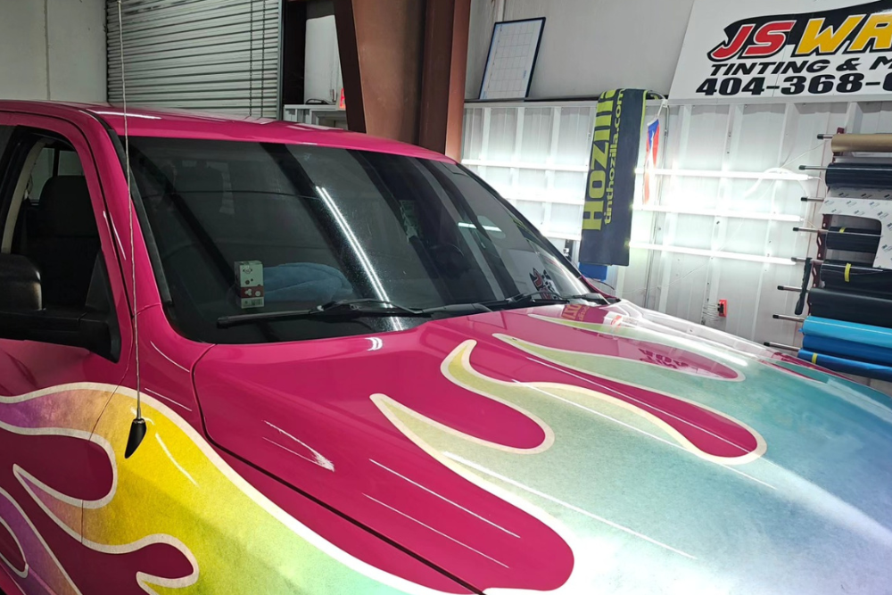 A pink truck with flames painted on the hood is parked in a garage.